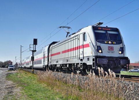 Deutsche Bahn locomotive pulling double-decker passenger cars along tracks with overhead wires against a blue sky, rural setting.