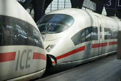 Two white ICE high-speed trains with red stripes at a station platform, front cockpits and windows visible, reflecting surrounding structure.